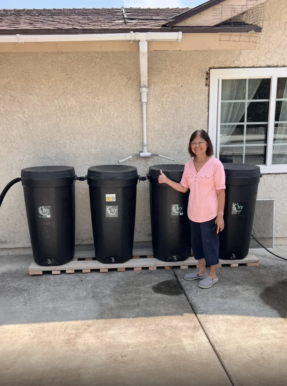 Easy Break Service customer in San Diego standing beside her newly installed rain‑barrel collection system on a scrap‑wood stand